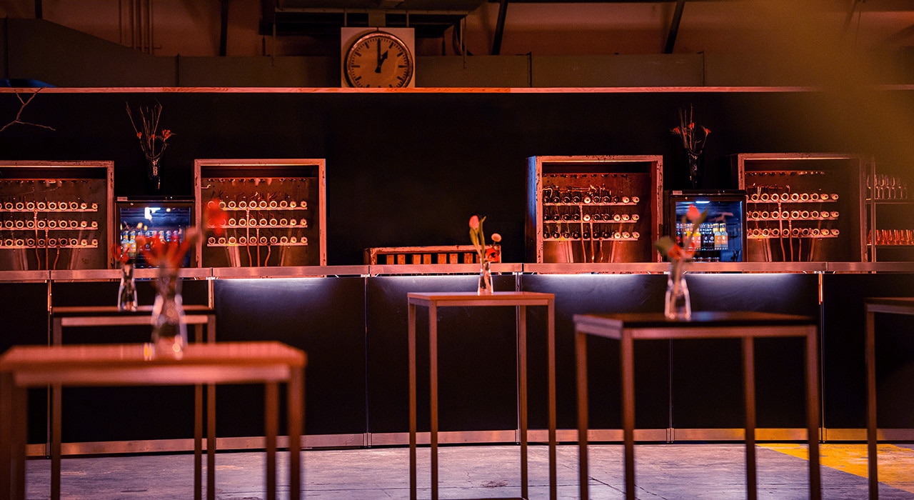 Die Bar unserer Eventlocation Leipzig, der Alten Wollkämmerei, wird hier dämmrig beleuchtet im abendlichen Ambiente gezeigt. Sie verleiht jedem Event einen modernen industriellen Charme.