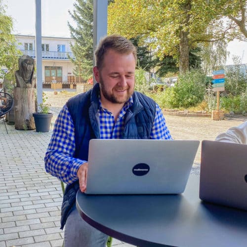 Unser Geschäftsführer sitzt draußen auf der Terrasse vor dem Büro und arbeitet lächelnd an einem Mac Book.