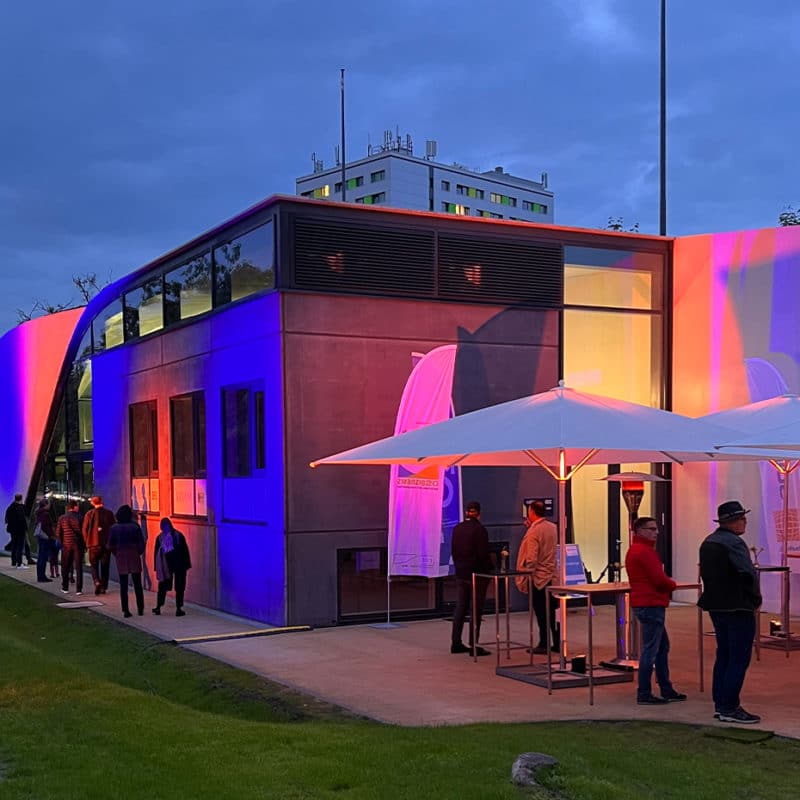 Eröffnungsfeier des Carbon Cubes. Farbenfroh beleuchtetes Haus bei Abenddämmerung. Auf dem Vorplatz unterhalten sich die Gäste an Stehtischen.